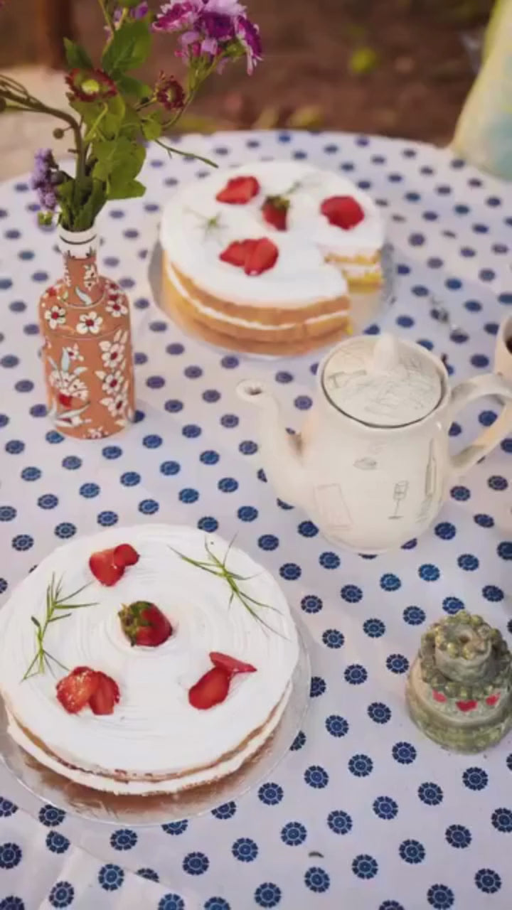 Woman in a light blue embroidered outfit standing outdoors with flowers and a white curtain in the background