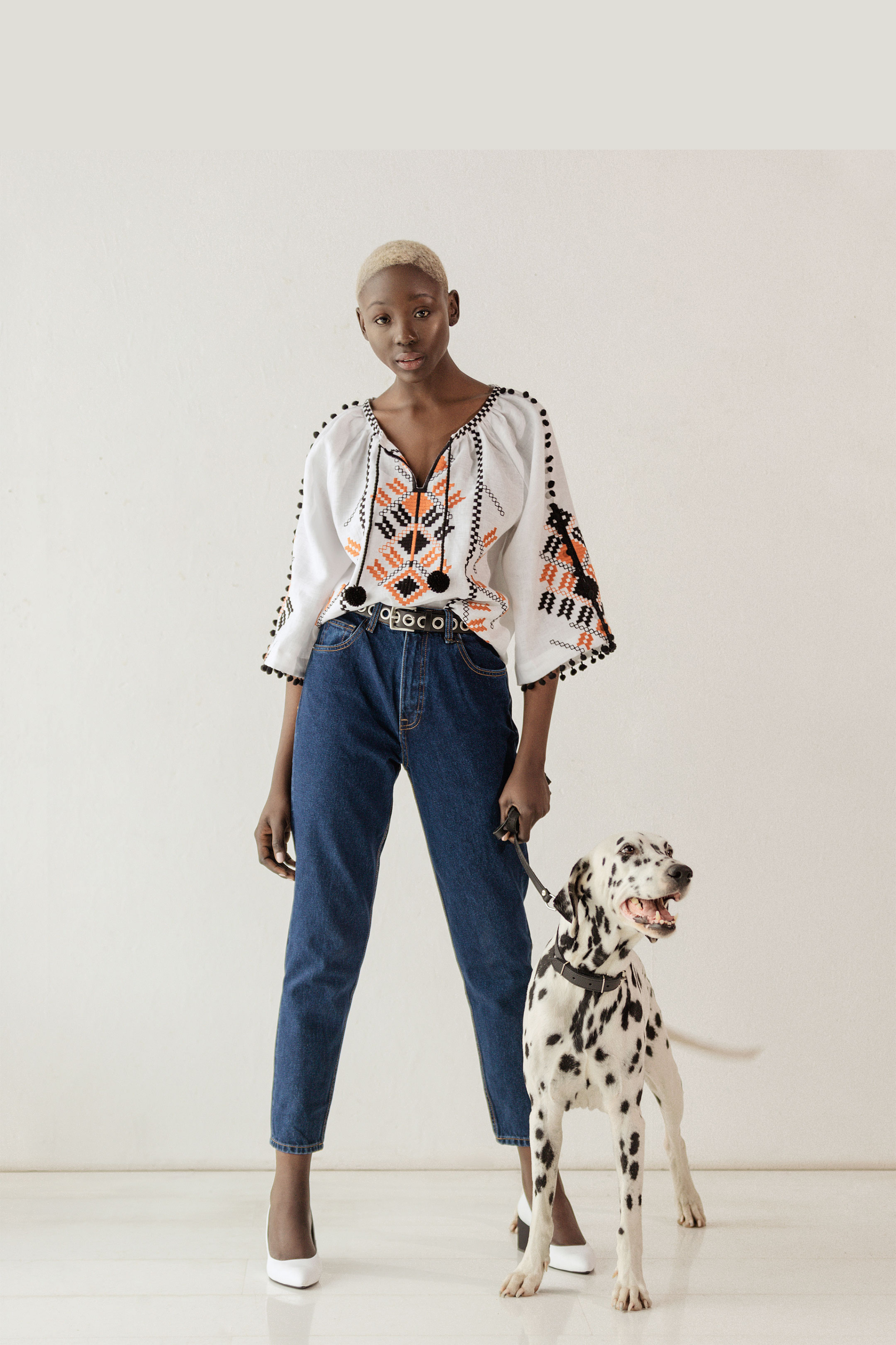 Woman in a patterned blouse and jeans standing next to a Dalmatian dog on a white background