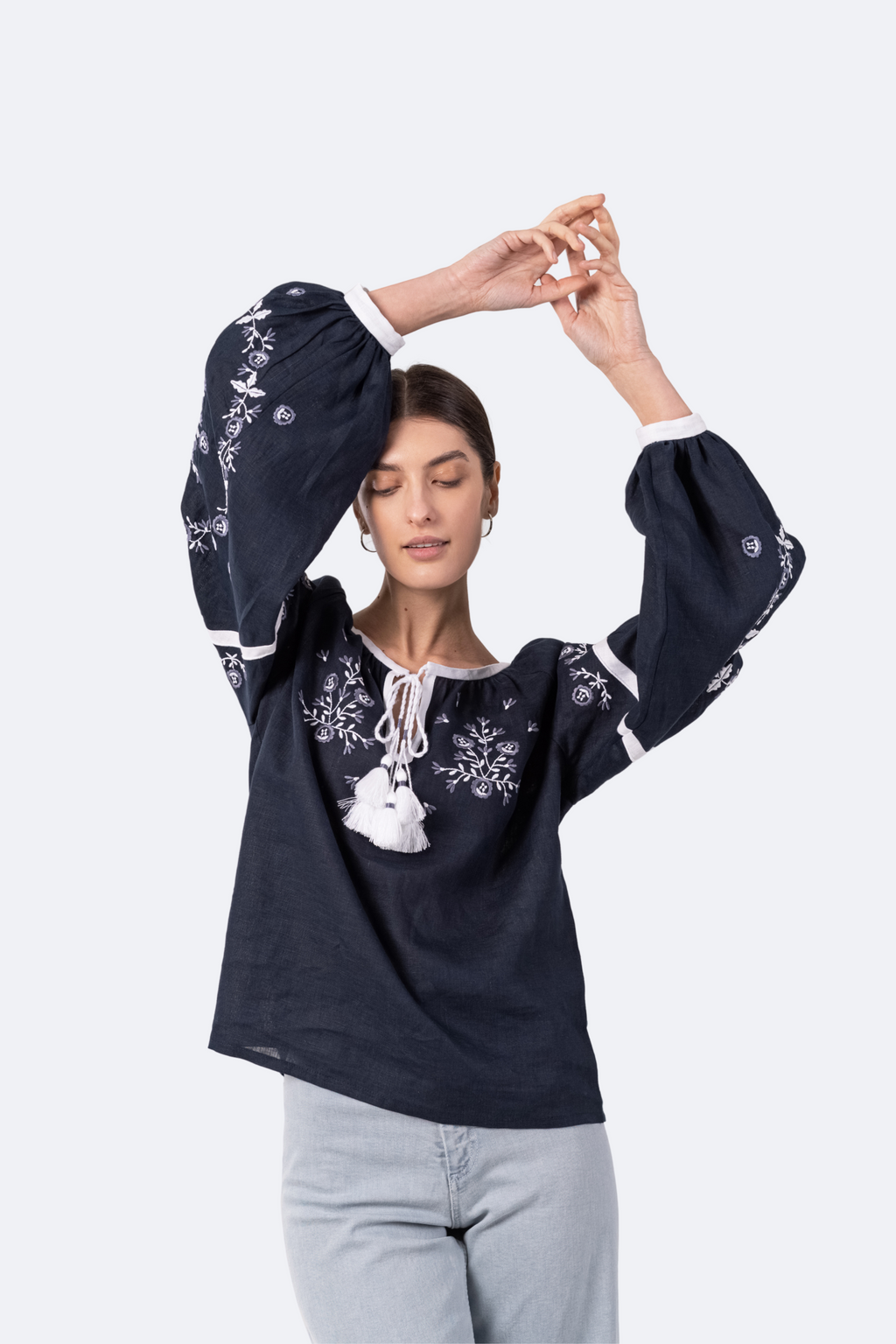 Woman wearing a dark blue embroidered blouse with white pants on a light gray background